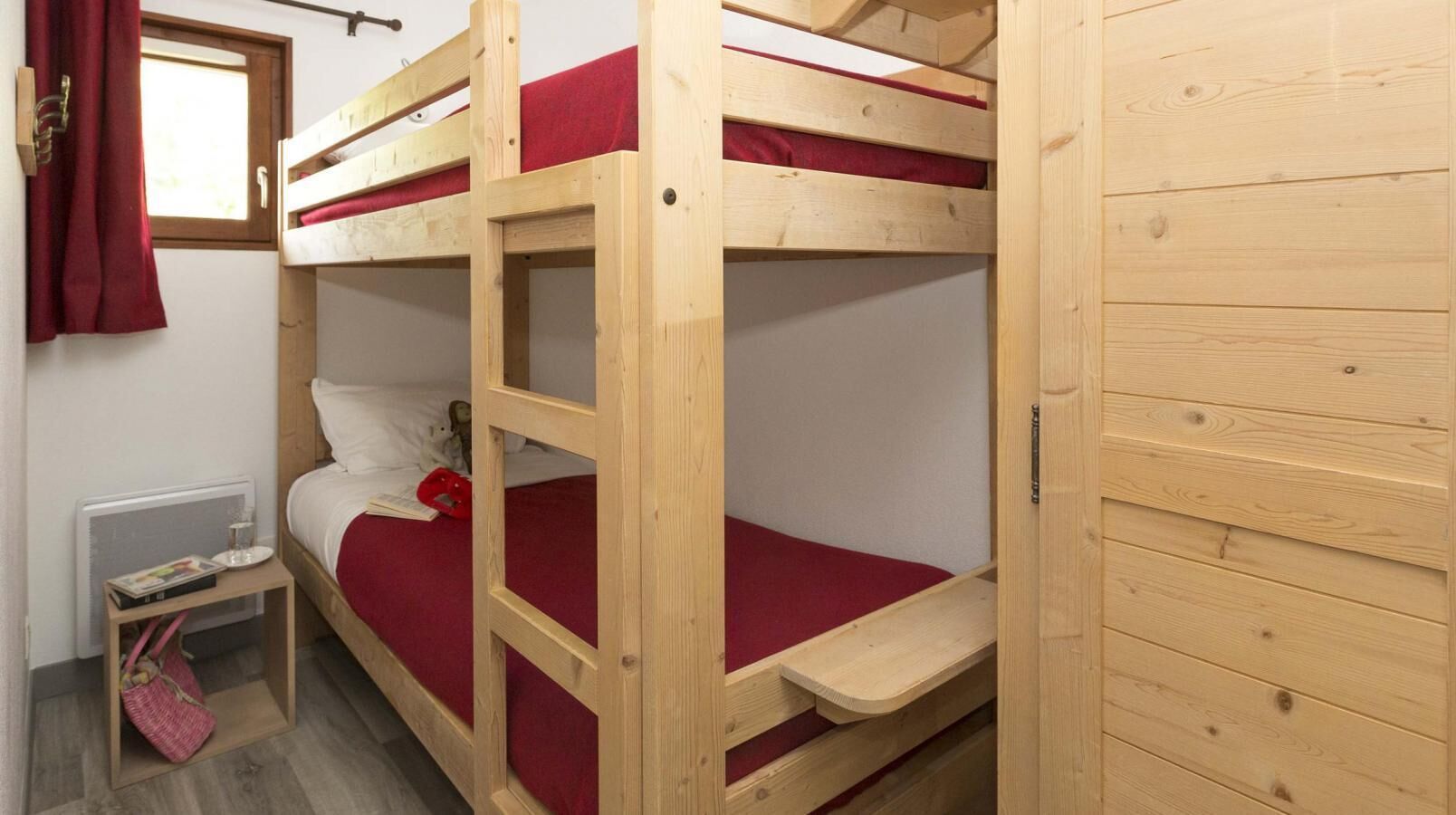 A bunk bed room with a red bedspread, a small bedside table with a book and a red object, and a window with a curtain.
