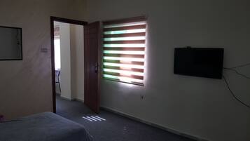 Una habitación con una cama, un televisor en la pared, una ventana con persianas y una puerta que da a otra estancia.