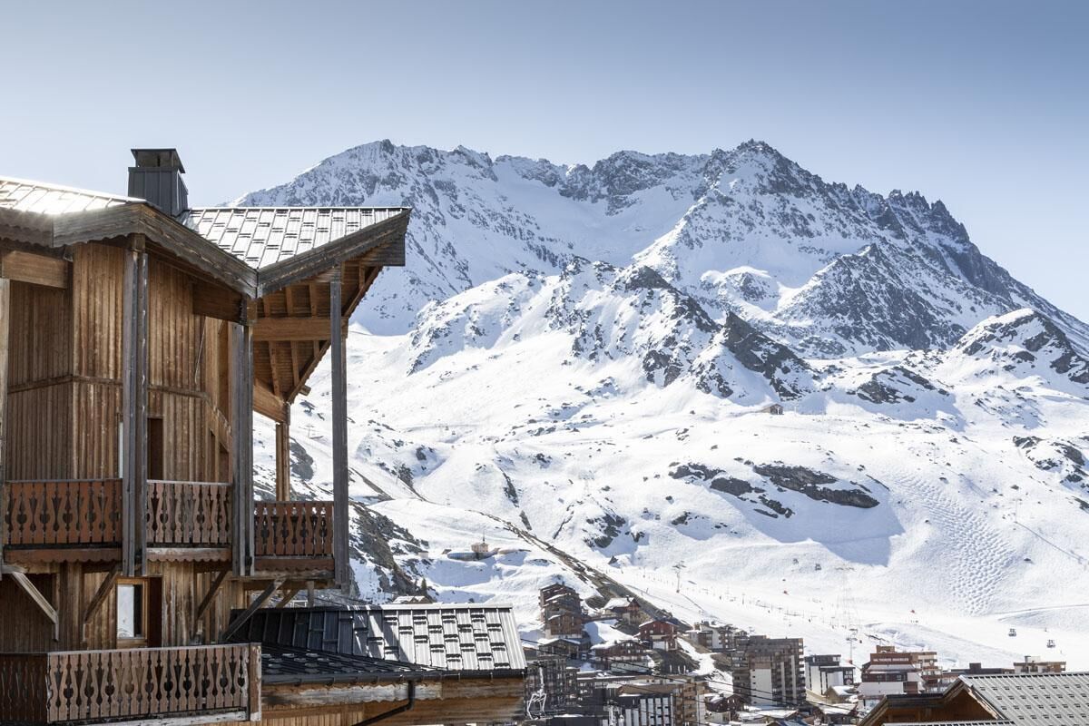 Una cabaña de madera con balcón, con montañas nevadas y un telesilla de fondo.