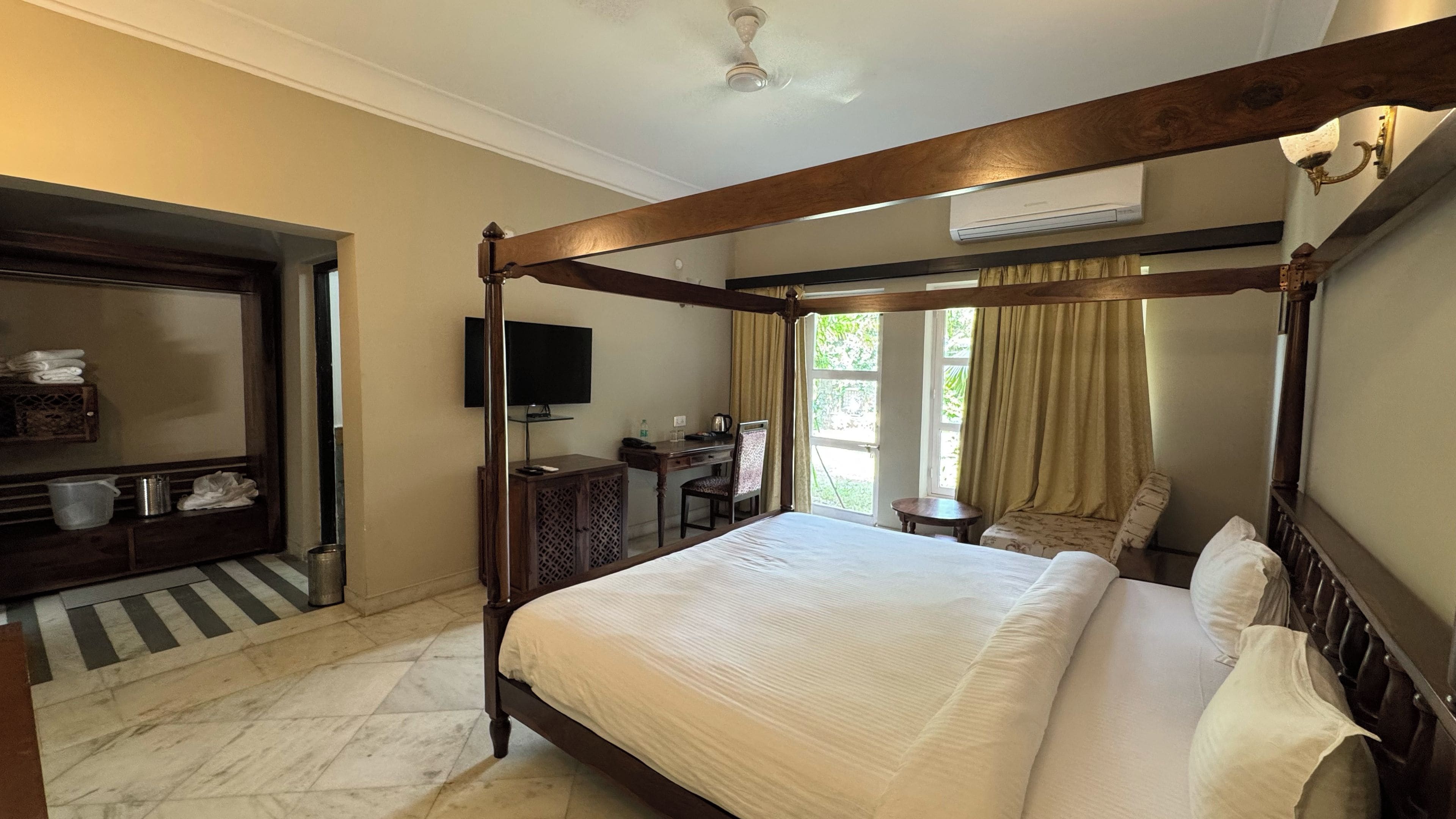A four-poster bed with a wooden frame, a flat-screen TV mounted on the wall, a desk with a chair, and a window with curtains.
