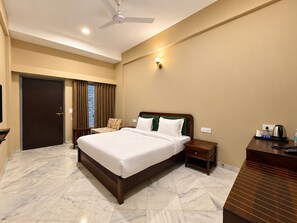 A hotel room with a bed, a desk, a chair, a nightstand, a ceiling fan, and a window with curtains. - King’s Abode - Luxuria Ranakpur by Moustache (Desuri)