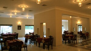 A dining area with multiple tables and chairs, ceiling lights, and large windows allowing natural light. - King's Abode Ranakpur (Desuri)