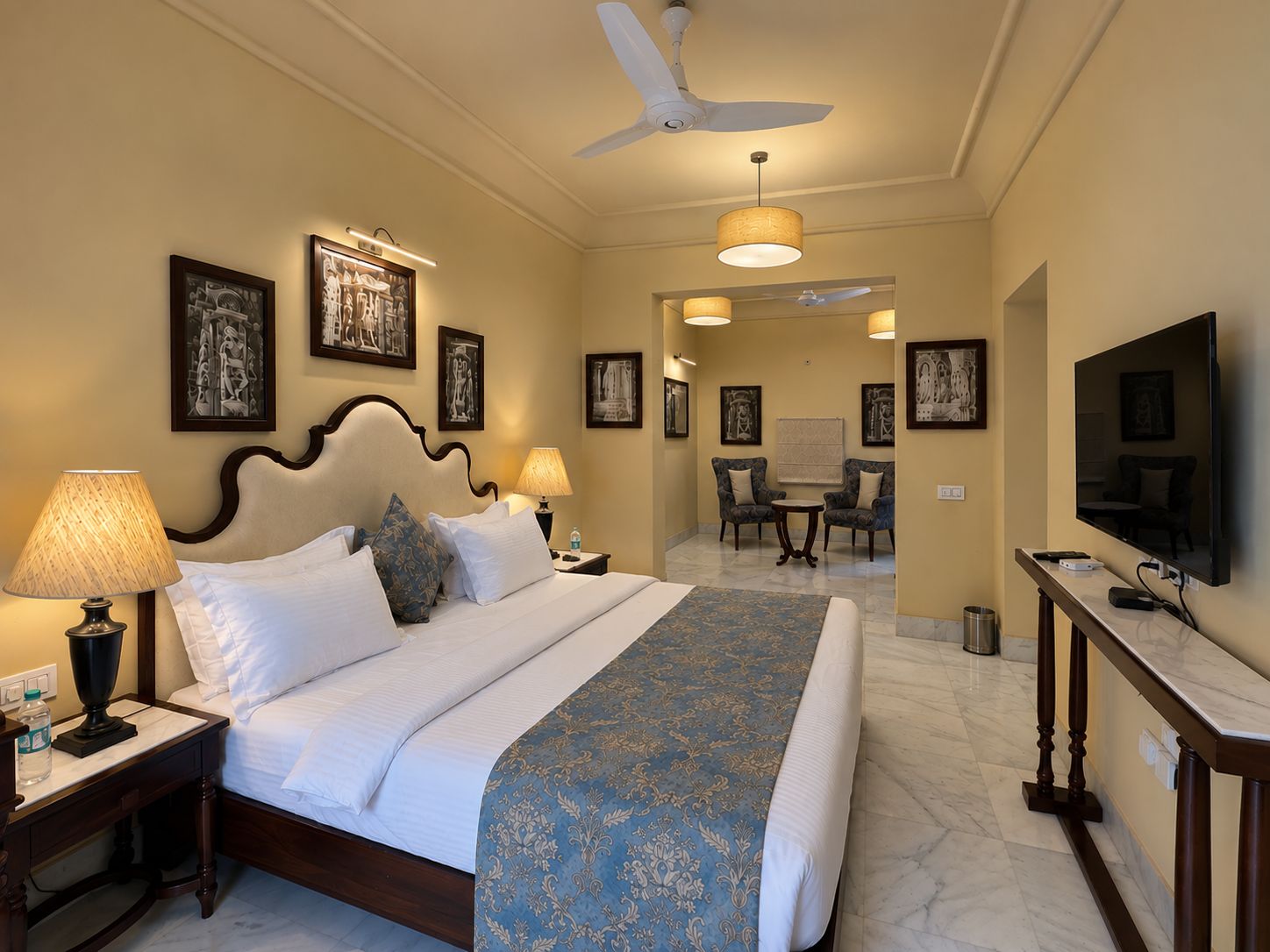 A hotel room with a bed, bedside lamps, a ceiling fan, framed artwork on the walls, a television, and a desk with a chair.