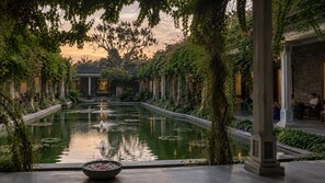 Une cour paisible, agrémentée d’une fontaine centrale, entourée d’une végétation luxuriante et bordée d’édifices à l’architecture traditionnelle.