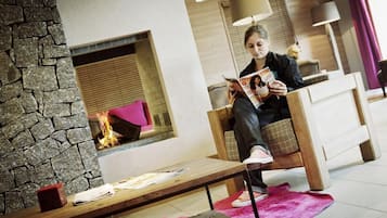 Une personne est assise sur une chaise en bois, lisant un magazine dans une pièce dont le mur est en pierre, qui contient une cheminée et est agrémentée d’un tapis rose.