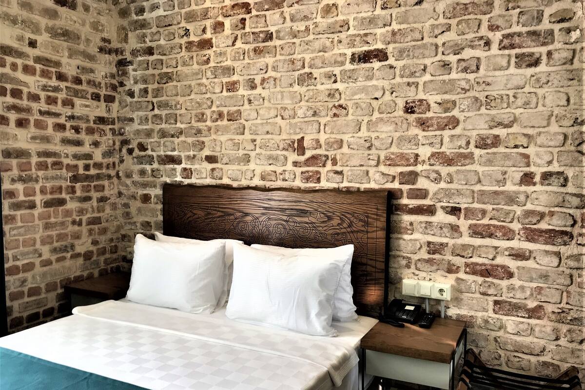 A room with a brick wall, a wooden headboard, a bed with white bedding, a bedside table with a phone, and a bench.