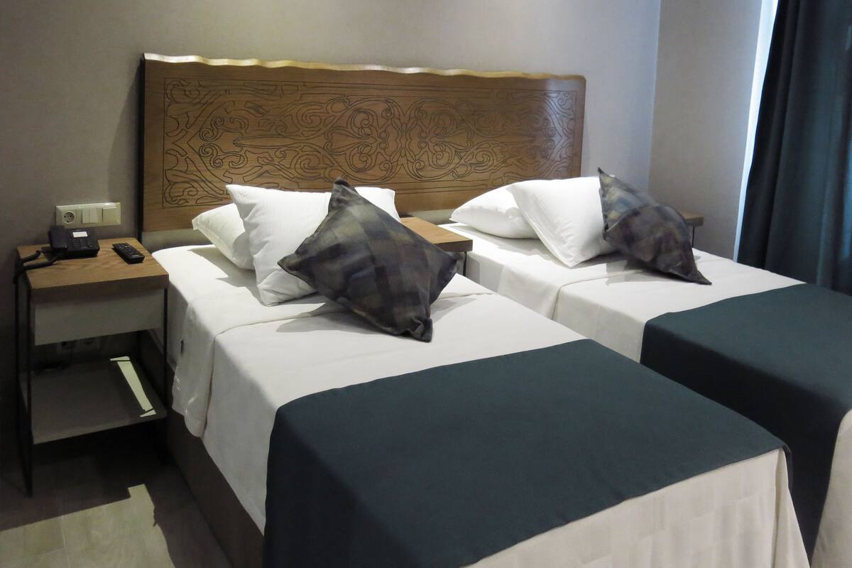 A hotel room with two beds, each with a pillow and a blanket. There is a nightstand with a phone and a lamp.