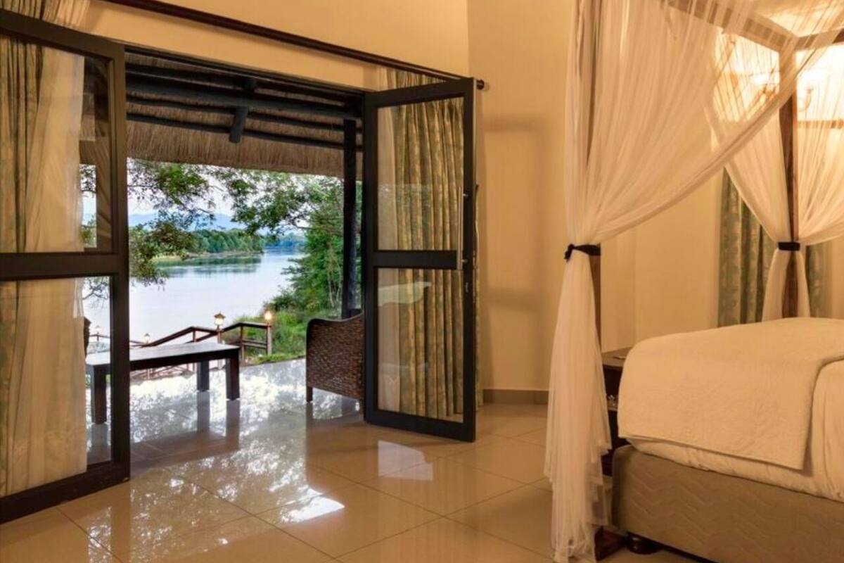 A room with a bed, a view of a lake, and a wooden bench outside.