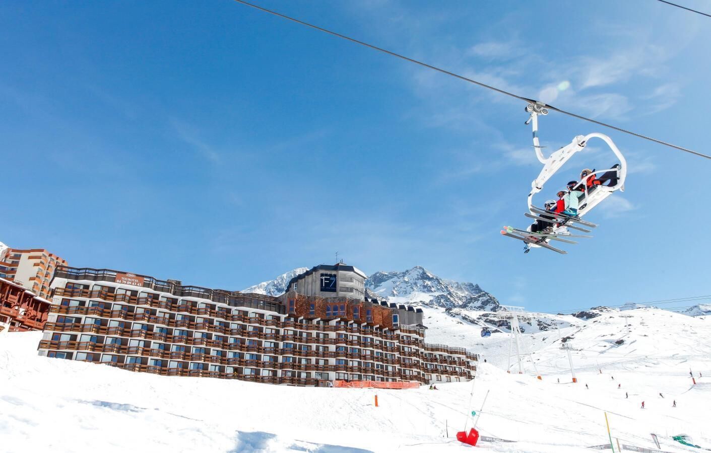 A ski lift with skiers, a hotel with multiple balconies, and snow-covered mountains in the background.