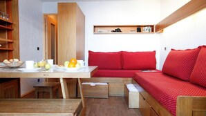 A compact room with a wooden dining table, a bench, a red sofa with storage drawers, and a shelf with books and a camera. - 300m from resort center, balcony, tv, ski locker, 16m², Val Thorens (Val Thorens)