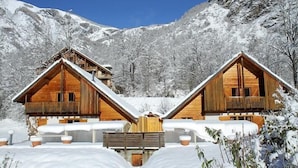 Deux cabanes en bois aux toits inclinés, entourées de montagnes enneigées.