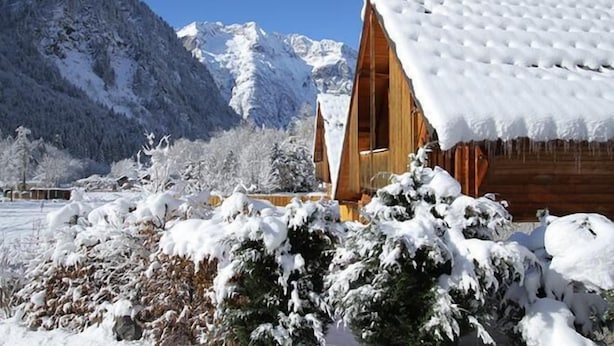 Une cabane en bois, dont le toit est recouvert de neige, entourée d’arbres et de montagnes enneigés.