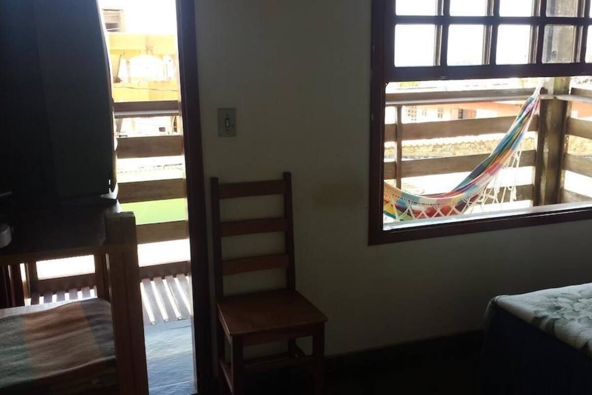 A room with a bed, a chair, a small table, and a window with a hammock outside.