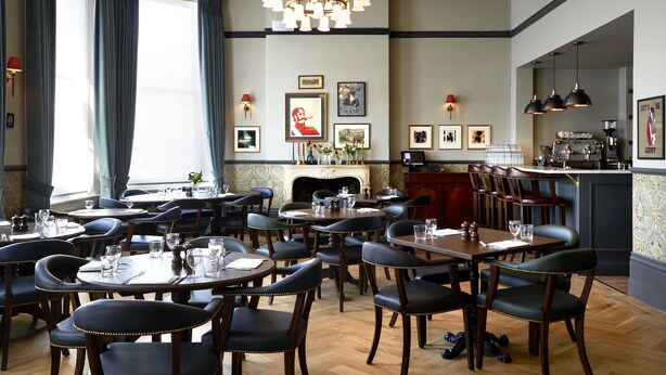 A formal dining room with multiple tables and chairs, a chandelier, framed pictures on the wall, and a fireplace. - The Hero of Maida (London)