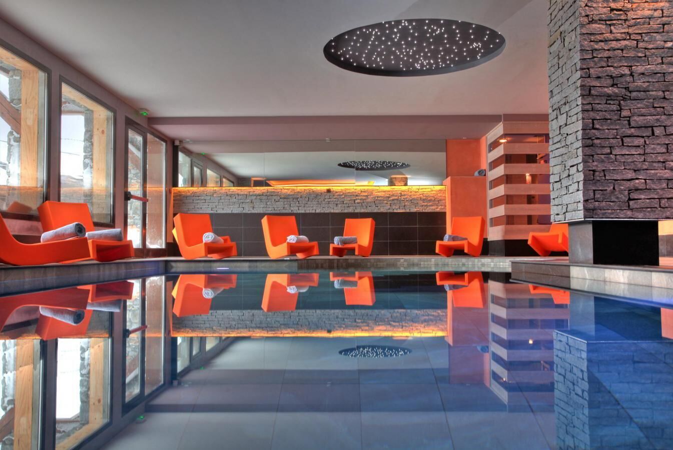 A modern indoor pool area with clear glass walls, orange lounge chairs, and a stone accent wall.