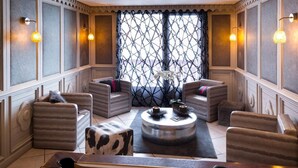A pool table with billiard balls, a pool cue, and a billiard rack. There are four cushioned chairs around a central coffee table. The room has a patterned window and wall paneling.