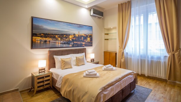 A hotel room with a large bed, a nightstand with a lamp, a painting of a cityscape, and a window with curtains. - Feel Belgrade Superior Apartment (Belgrade)