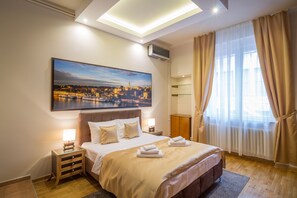 A hotel room with a large bed, a nightstand with a lamp, a painting of a cityscape, and a window with curtains.