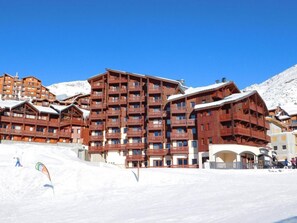 Um hotel com vários andares e varandas, uma pista de esqui em primeiro plano, e um céu azul límpido.