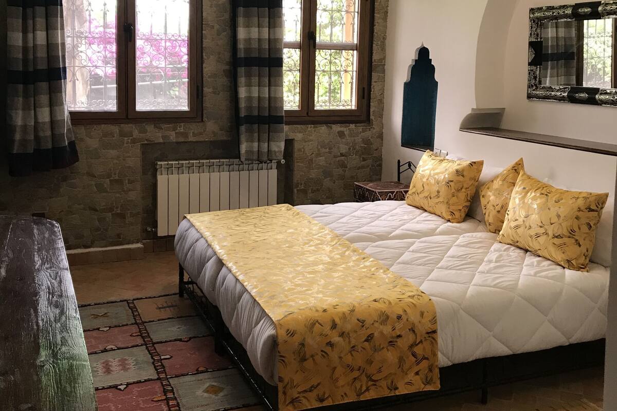 Ein Schlafzimmer mit einem großen Bett, zwei Fenstern mit Vorhängen, einem Heizkörper und einem gemusterten Teppich.