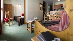 Een compacte kamer met een bed, een klein tafeltje, een stoel, een houten garderobe en een kitchenette.