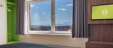 A hotel room with a bed, a window with a view of mountains, a green wall, and a television on the wall.