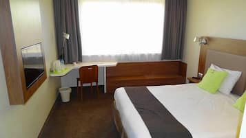 Ein Hotelzimmer mit Bett, Schreibtisch, Stuhl, Fernseher und einem Fenster mit Vorhängen.