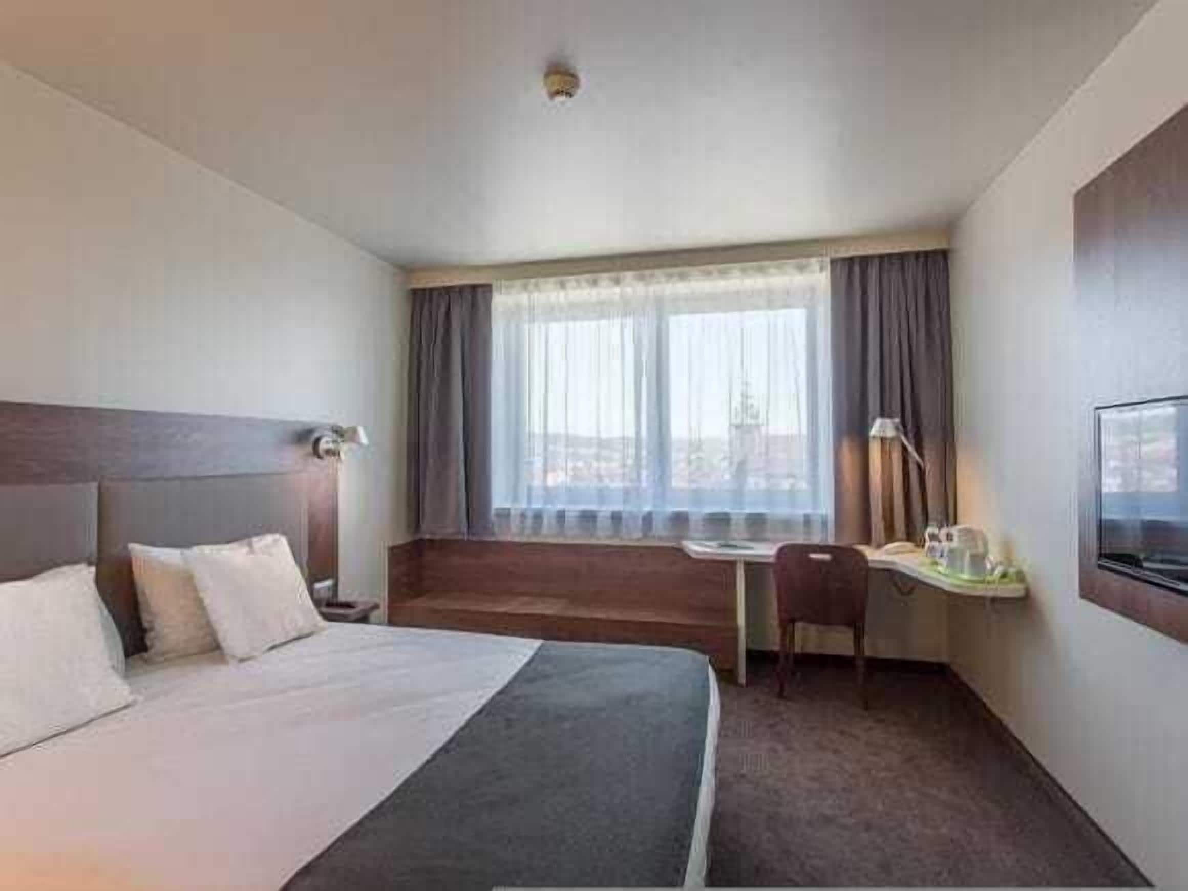 Ein Hotelzimmer mit Bett, Schreibtisch, Stuhl, Fernseher und einem Fenster mit Vorhängen.