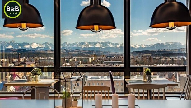 Une chambre d’hôtel dotée de grandes fenêtres offrant une vue sur des montagnes enneigées et sur le paysage urbain. Trois lampes suspendues sont accrochées au-dessus d’une table ornée de bougies et d’une plante en pot.