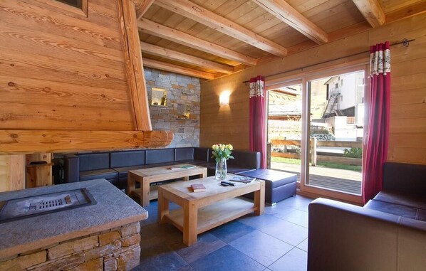 A cozy interior with a stone fireplace, wooden beams, and a stone wall. There are seating areas with cushions and a coffee table.