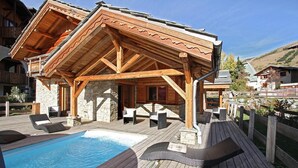 A wooden deck with a pool, lounge chairs, and a covered seating area with stone walls. - Chalet, 50m from resort center, swimming pool, sauna, balcony, fireplace or stove, parking, tv (Les Deux Alpes)