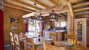 A wooden cabin interior with a dining area, kitchen, and a large window. - Chalet, 50m from resort center, swimming pool, sauna, balcony, fireplace or stove, parking, tv (Les Deux Alpes)