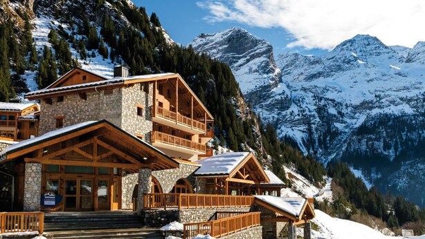 Un chalet de montagne construit en pierre et en bois, doté de plusieurs balcons et offrant une vue sur des montagnes enneigées.