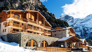 Un chalet en bois à plusieurs étages, doté de fondations en pierre, se dressant sur un fond de montagnes enneigées.