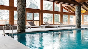 Une piscine intérieure agrémentée de piliers en pierre, de poutres en bois et de grandes fenêtres offrant une vue sur un paysage montagneux.