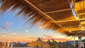 Un restaurante en la playa con mesas y sillas de madera, sombrillas de paja y vistas al océano al atardecer.