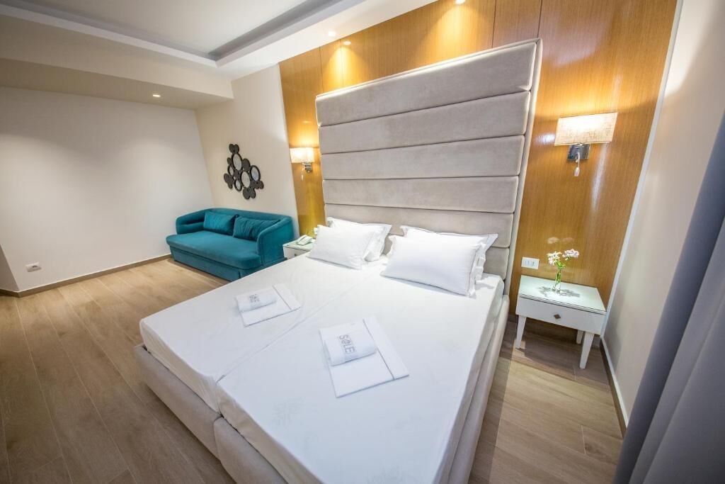 A hotel room with a large bed, a teal sofa, a nightstand with a lamp, and a wall-mounted artwork.