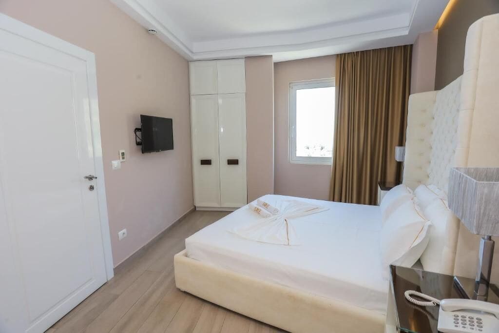 A hotel room with a bed, a bedside telephone, a TV mounted on the wall, a wardrobe, and a window with curtains.