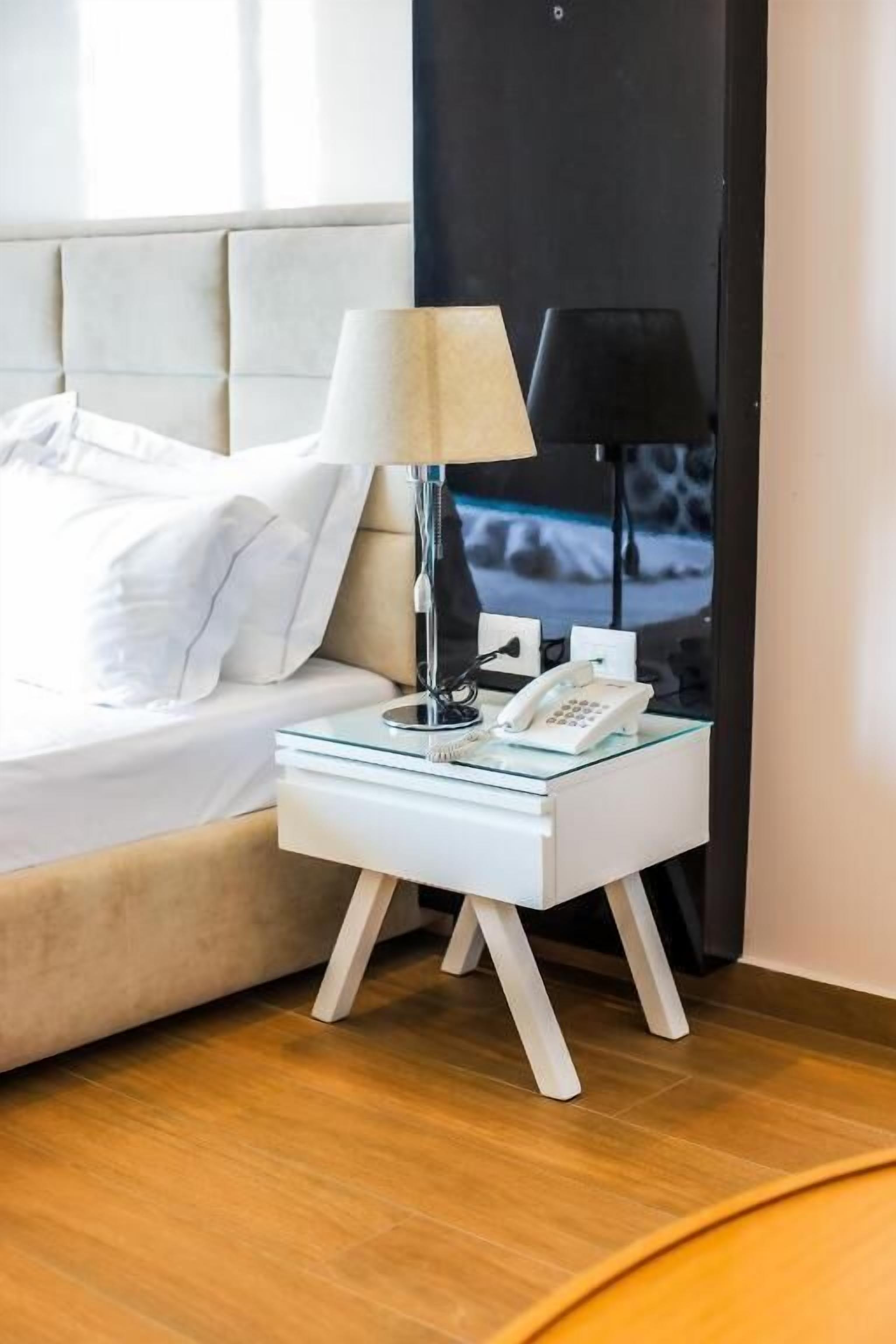 Un letto rifatto con lenzuola bianche, un tavolino in vetro con una lampada, un telefono e un oggetto nero sul tavolo.