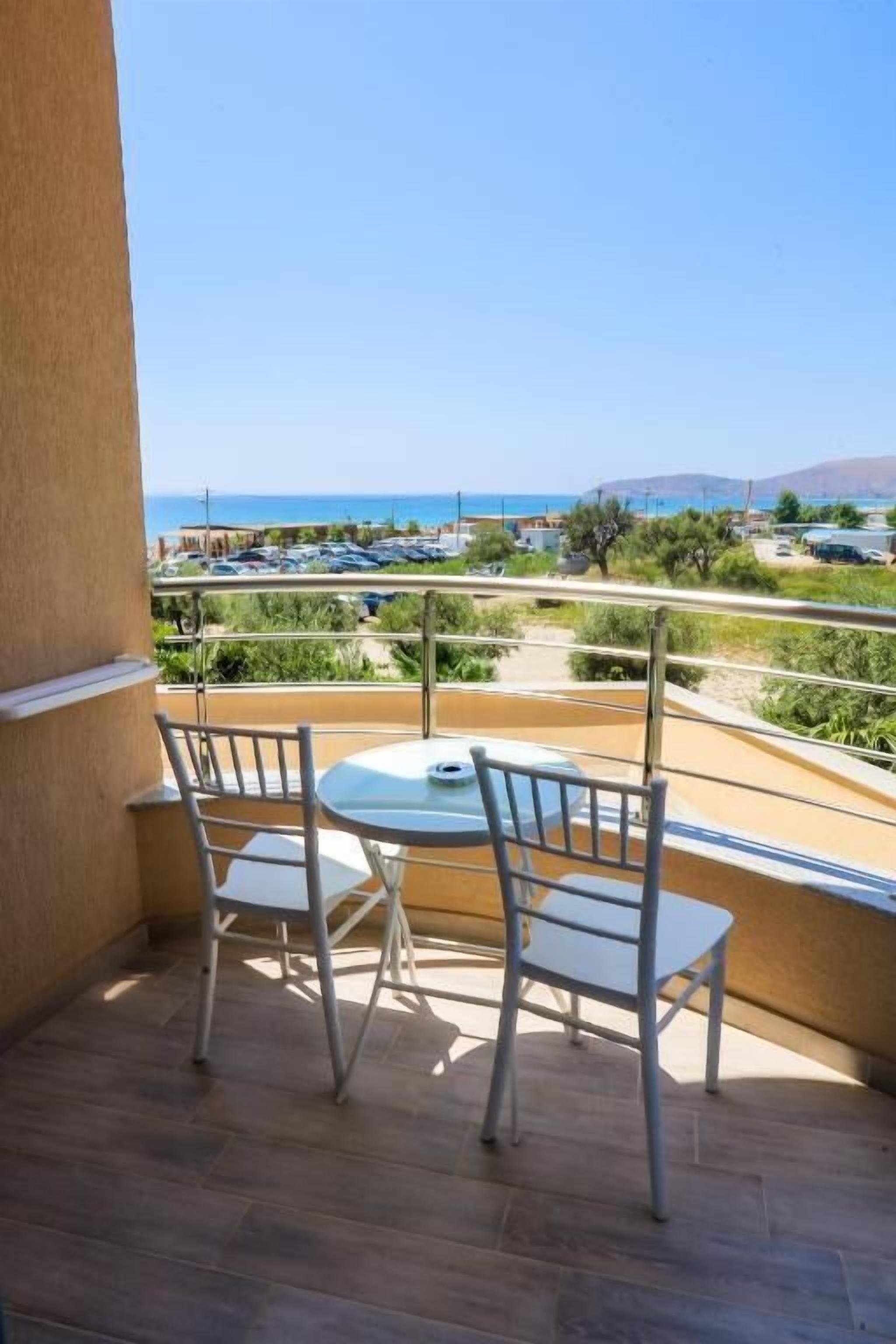 A balcony with a table and two chairs, overlooking a coastal area with parked cars and greenery.