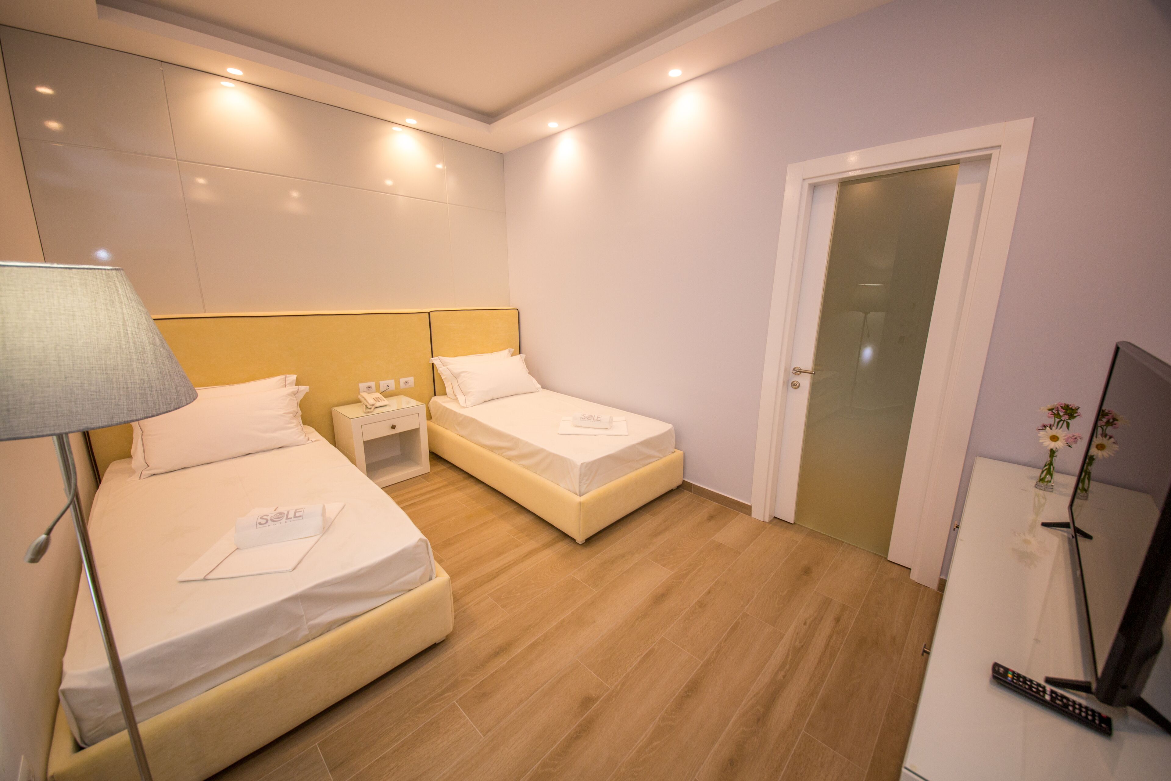 A hotel room with two beds, a nightstand, a lamp, a TV, and a door.
