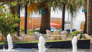 Una fuente con chorros de agua, rodeada de una zona pavimentada con sillas y mesas, y árboles que dan sombra.