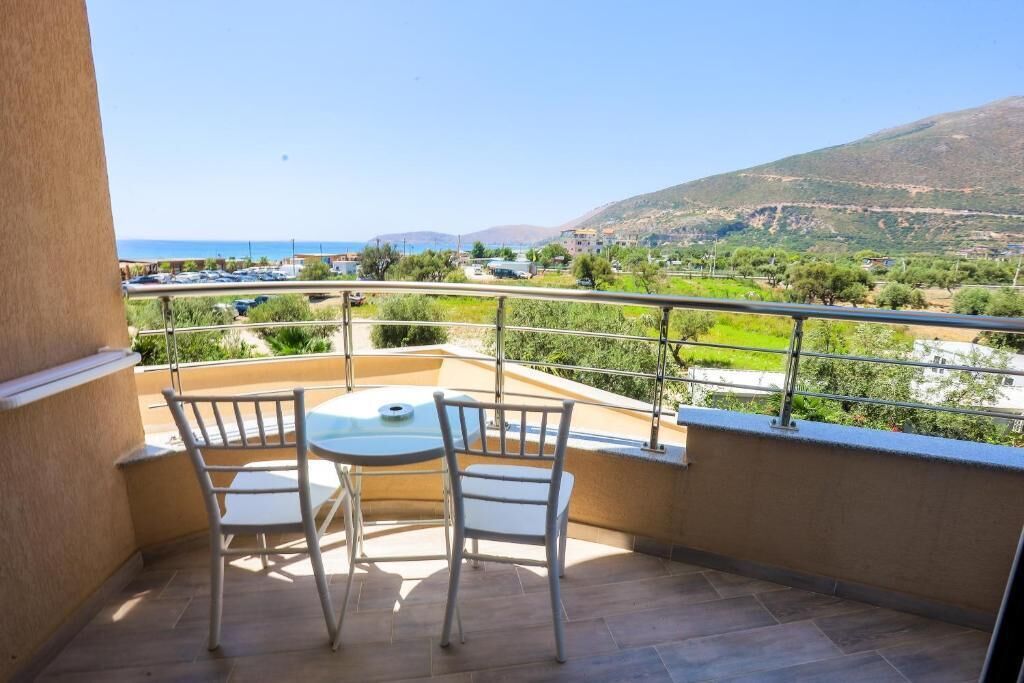 A balcony with a table and two chairs, overlooking a coastal landscape with buildings and hills.