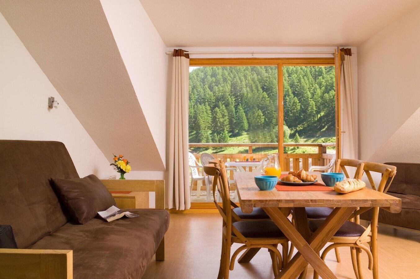 A cozy room with a wooden dining table set for breakfast, a brown sofa, and a view of a forest through large windows.