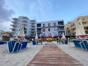 A beachside hotel with a wooden walkway leading to the entrance, surrounded by beach umbrellas and lounge chairs.