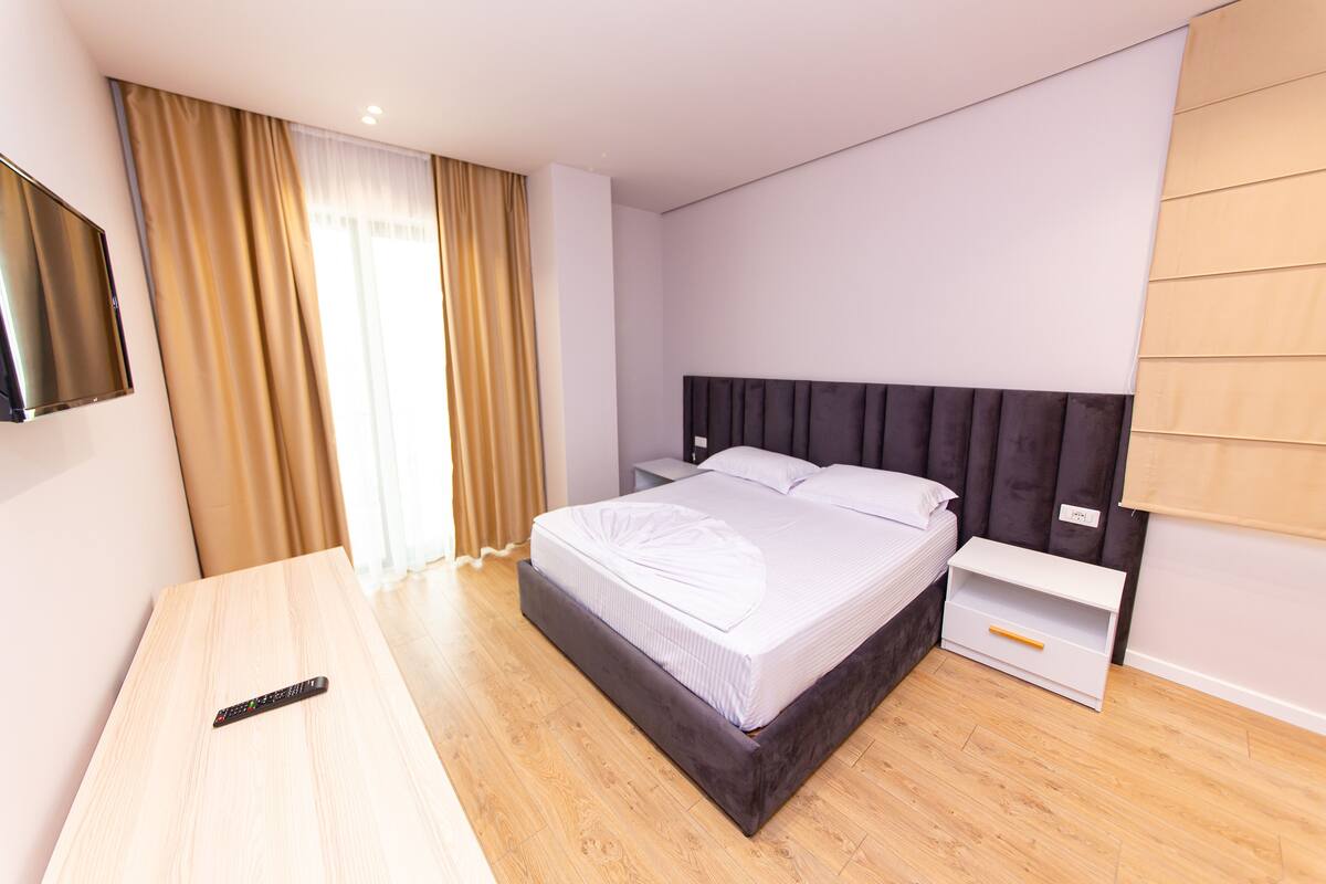 A modern hotel room with a large bed, a bedside table, a TV mounted on the wall, and a window with curtains.