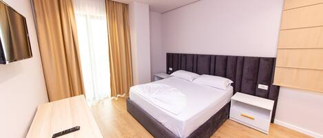 A modern hotel room with a large bed, a bedside table, a TV mounted on the wall, and a window with curtains.