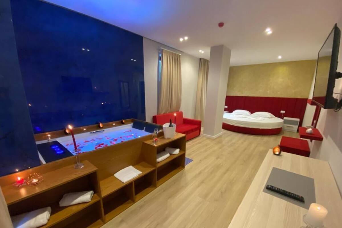 A modern hotel room with a large bed, a small seating area, a desk, and a large window.