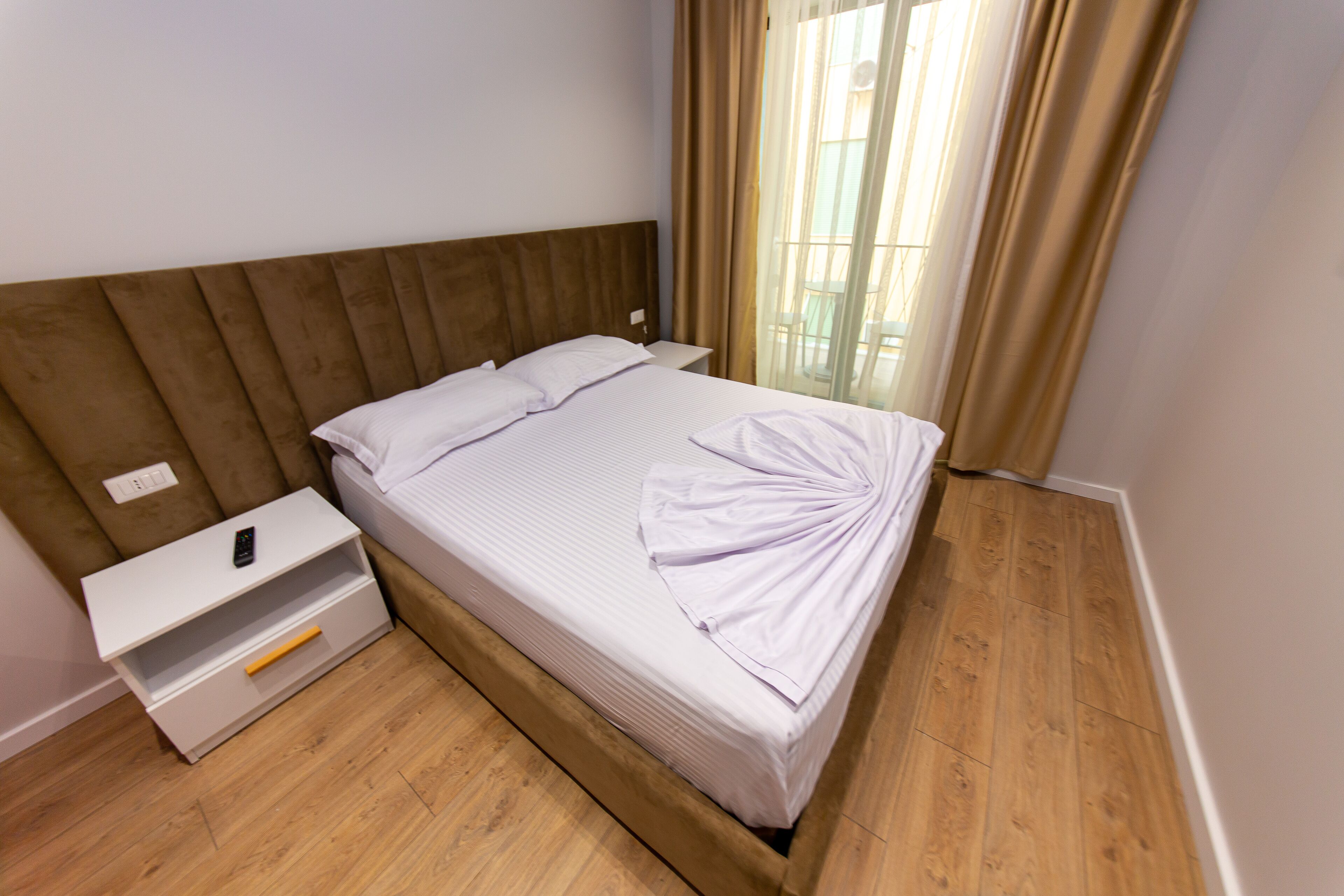A neatly made bed with a wooden headboard, a white bedside table with a drawer, and a window with curtains.