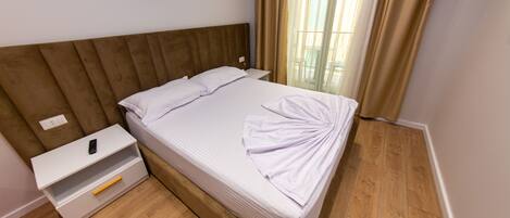A neatly made bed with a wooden headboard, a white bedside table with a drawer, and a window with curtains.